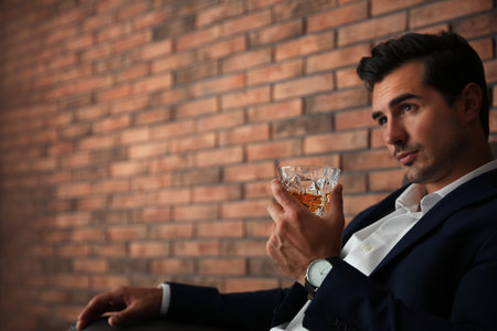 Young man with glass of whiskey near brick wall indoors. Space for textの写真素材
