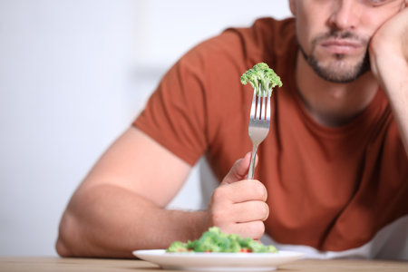 Unhappy man with broccoli on fork at table, closeupの写真素材