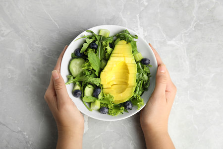 Woman holding bowl of avocado salad with blueberries over gray marble table, top view
