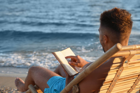 Young man reading book on sandy beach near seaの写真素材