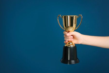 Child holding golden winning cup on dark blue background, closeup. Space for textの写真素材