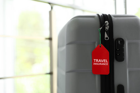 Stylish suitcase with travel insurance label on blurred background, closeup. Space for textの写真素材