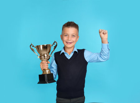 Happy boy in school uniform with golden winning cup on blue background. Space for textの写真素材