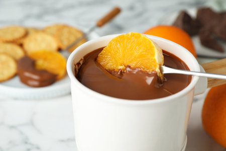 Dipping orange into fondue pot with chocolate on white marble table, closeupの写真素材