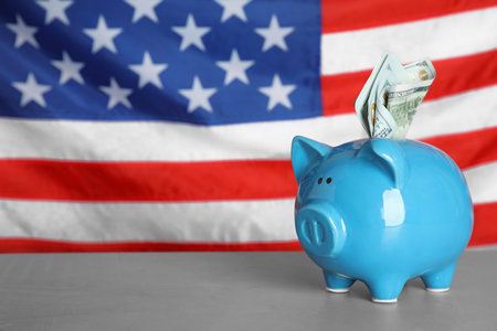 Blue piggy bank with banknotes on light gray table against national USA flag. Space for textの写真素材