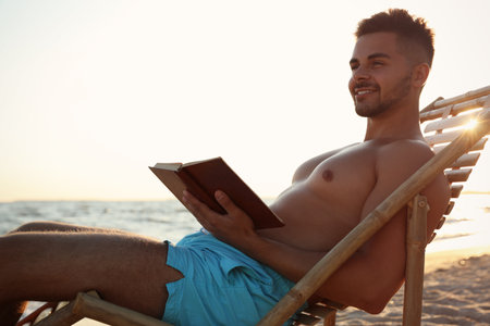 Young man reading book in deck chair on beachの写真素材