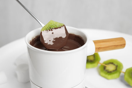 Dipping kiwi into fondue pot with chocolate on white tableの写真素材