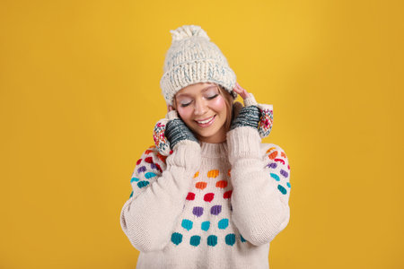 Young woman in warm sweater, mittens and hat on yellow background. Winter seasonの写真素材