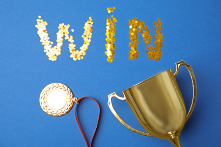 Gold trophy cup, medal and word WIN made with confetti on blue background, flat layの写真素材