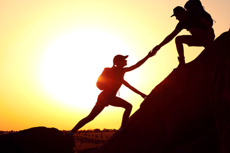 Silhouettes of man and woman helping each other to climb on hill against sunsetの写真素材
