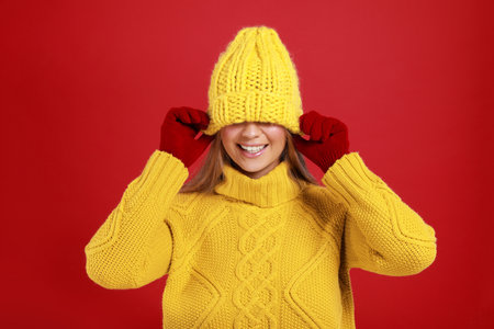 Funny young woman in warm sweater, gloves and hat on red background. winter season
