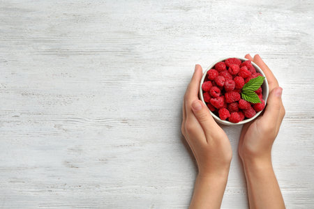 Woman holding bowl with delicious ripe raspberries on white wooden table, top view. Space for text
