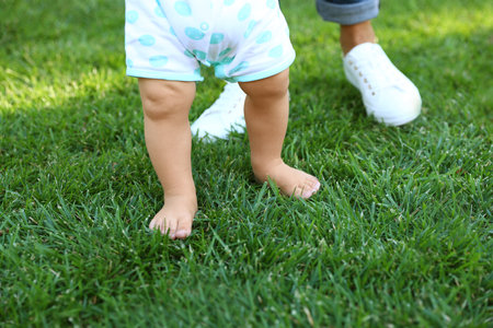 Cute little baby learning to walk with his nanny on green grass outdoors, closeupの写真素材