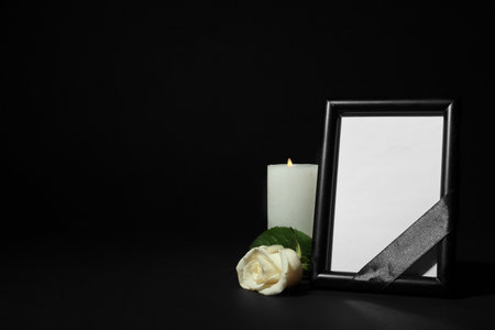 Funeral photo frame with ribbon, white rose and candle on dark table against black background. space for designの写真素材