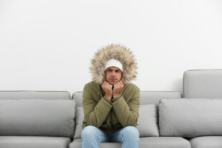 Young man in warm clothes freezing on sofa against white backgroundの写真素材