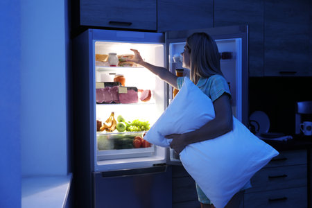 Woman with pillow looking into refrigerator in kitchen at nightの写真素材
