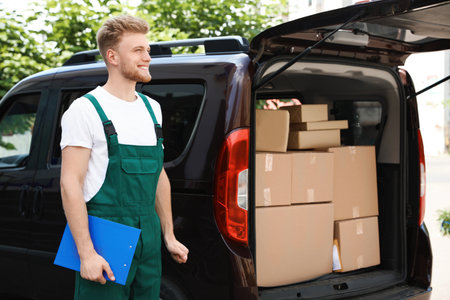 Young courier holding clipboard near delivery car with parcels outdoors