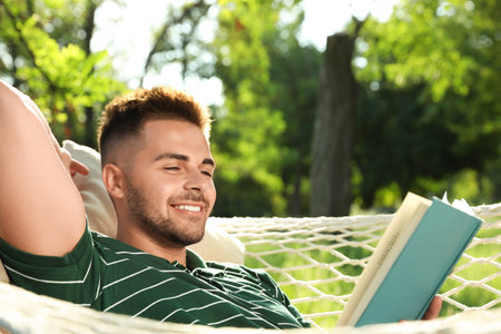 Young man reading book in comfortable hammock at green gardenの写真素材