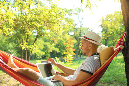 Young man with laptop resting in comfortable hammock at green gardenの写真素材