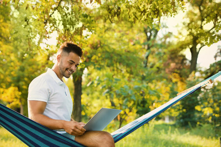 Young man with laptop in comfortable hammock at green gardenの写真素材