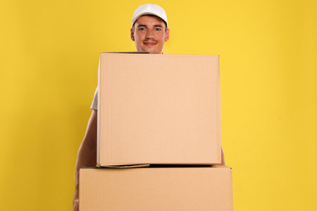 Happy young courier with cardboard boxes on yellow backgroundの写真素材