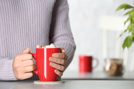 Woman holding cup of tasty cocoa with marshmallows at table indoors, closeup. Space for textの写真素材