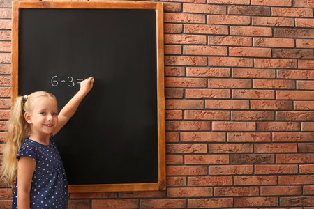 Cute little left-handed girl doing sums on chalkboard near brick wallの写真素材
