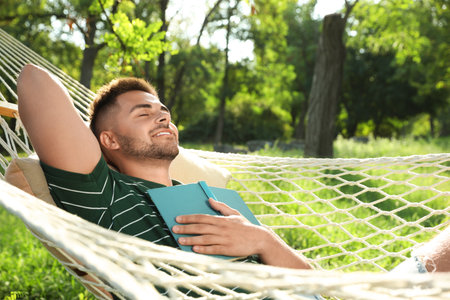 Young man with book resting in comfortable hammock at green gardenの写真素材