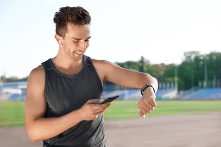 Young sportsman with wireless earphones, mobile phone and smart watch at stadiumの写真素材