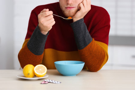 Sick young man eating soup to cure flu at table in kitchenの写真素材