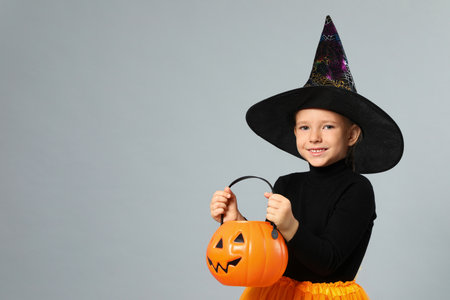 Cute little girl with pumpkin candy bucket wearing Halloween costume on gray background, space for...