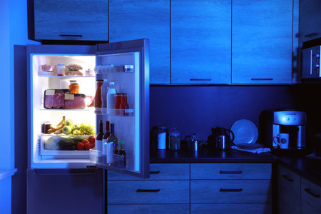 Open refrigerator full of products in kitchen at nightの写真素材