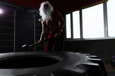 Young Santa Claus hitting heavy tire with hammer in modern gymの写真素材