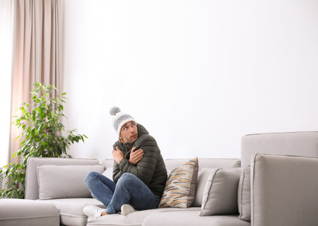 Young man wearing warm clothes freezing on sofa at home. air conditioner malfunctionの写真素材