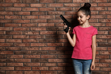 Little child playing with gun against brick wall, space for text. Dangerous gameの写真素材