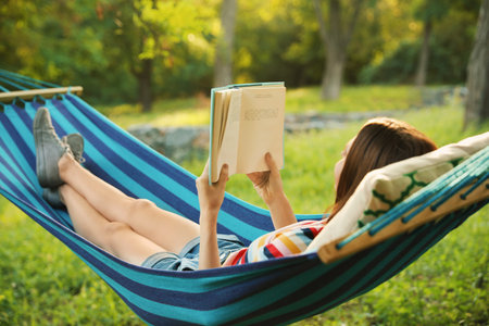 Young woman reading book in comfortable hammock at green gardenの写真素材