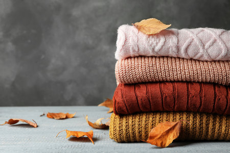 Stack of warm clothes and autumn leaves on wooden table against gray background. Space for textの写真素材