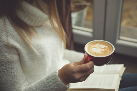 Woman with cup of coffee reading book near window indoors, closeupの写真素材