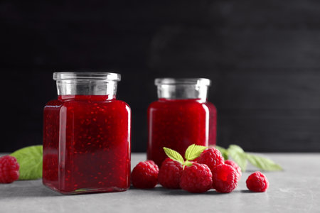 Glass jars of sweet jam with ripe raspberries and green leaves on table against black background....