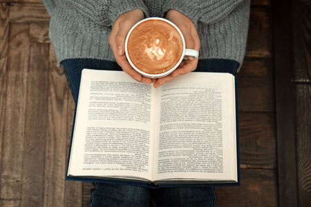 Woman with cup of coffee reading book at home, top view