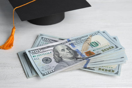 Dollar banknotes and student graduation hat on white wooden background. Tuition fees conceptの写真素材