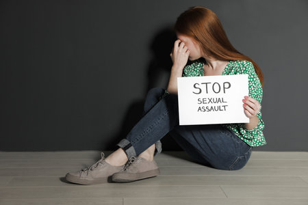 Young woman holding card with words STOP SEXUAL ASSAULT while sitting near gray wall. Space for textの写真素材