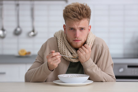 Sick young man eating soup to cure flu at table in kitchenの写真素材