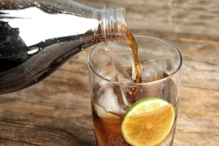 Pouring refreshing soda drink into glass with ice cubes and lime on wooden table, closeupの写真素材
