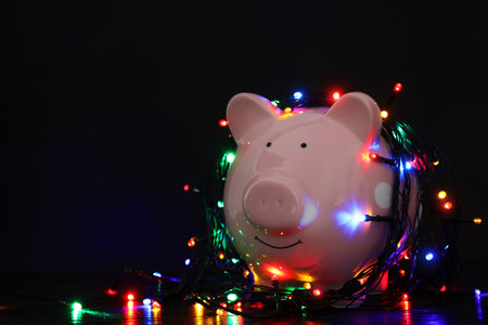 Piggy bank with Christmas lights on wooden table against black background. Space for textの写真素材