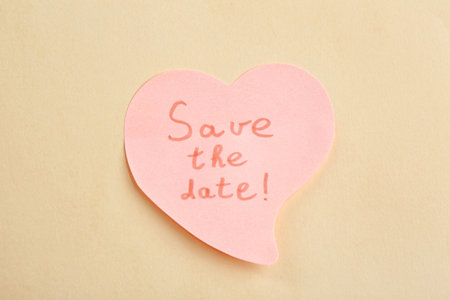 Heart shaped note with phrase SAVE THE DATE on beige background, top view