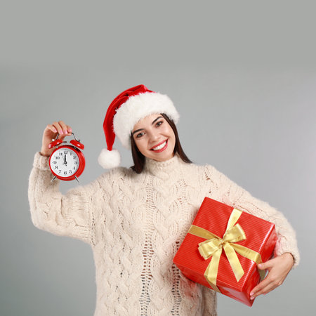 Happy young woman in Santa hat with Christmas gift and alarm clock on gray background