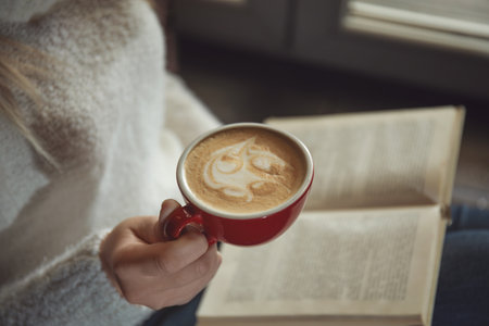 Woman with cup of coffee reading book near window indoors, closeupの写真素材