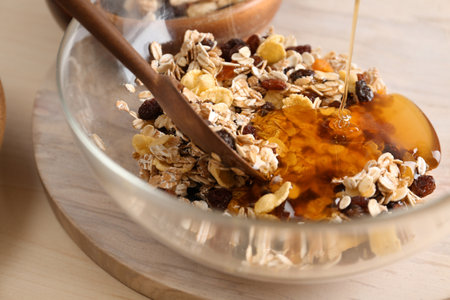 Pouring honey into bowl with oatmeal flakes and raisins on table, closeup. Healthy granola bar preparationの写真素材