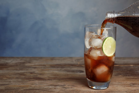 Pouring refreshing soda drink into glass with ice cubes and lime on wooden table against blue background. Space for textの写真素材
